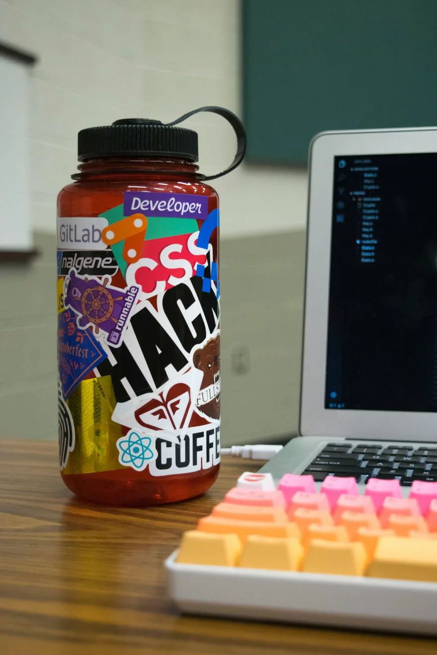 Stickers for water bottles on a red bottle next to a laptop and colorful keyboard.